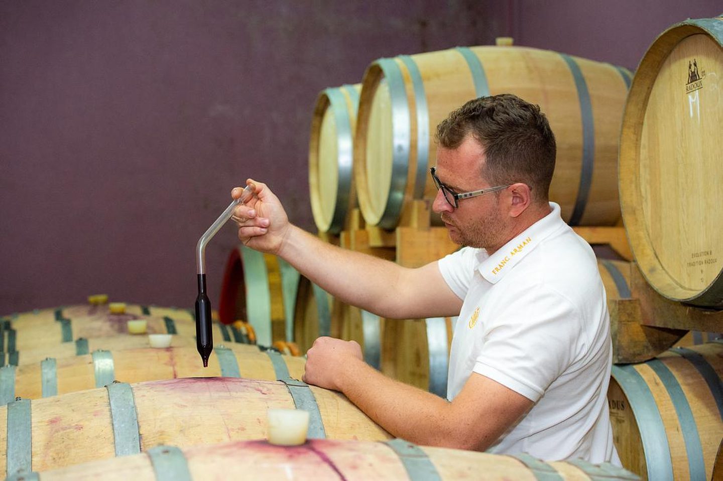 Winemaker in a Franc Arman polo drawing red wine from an oak barrique with a glass thief