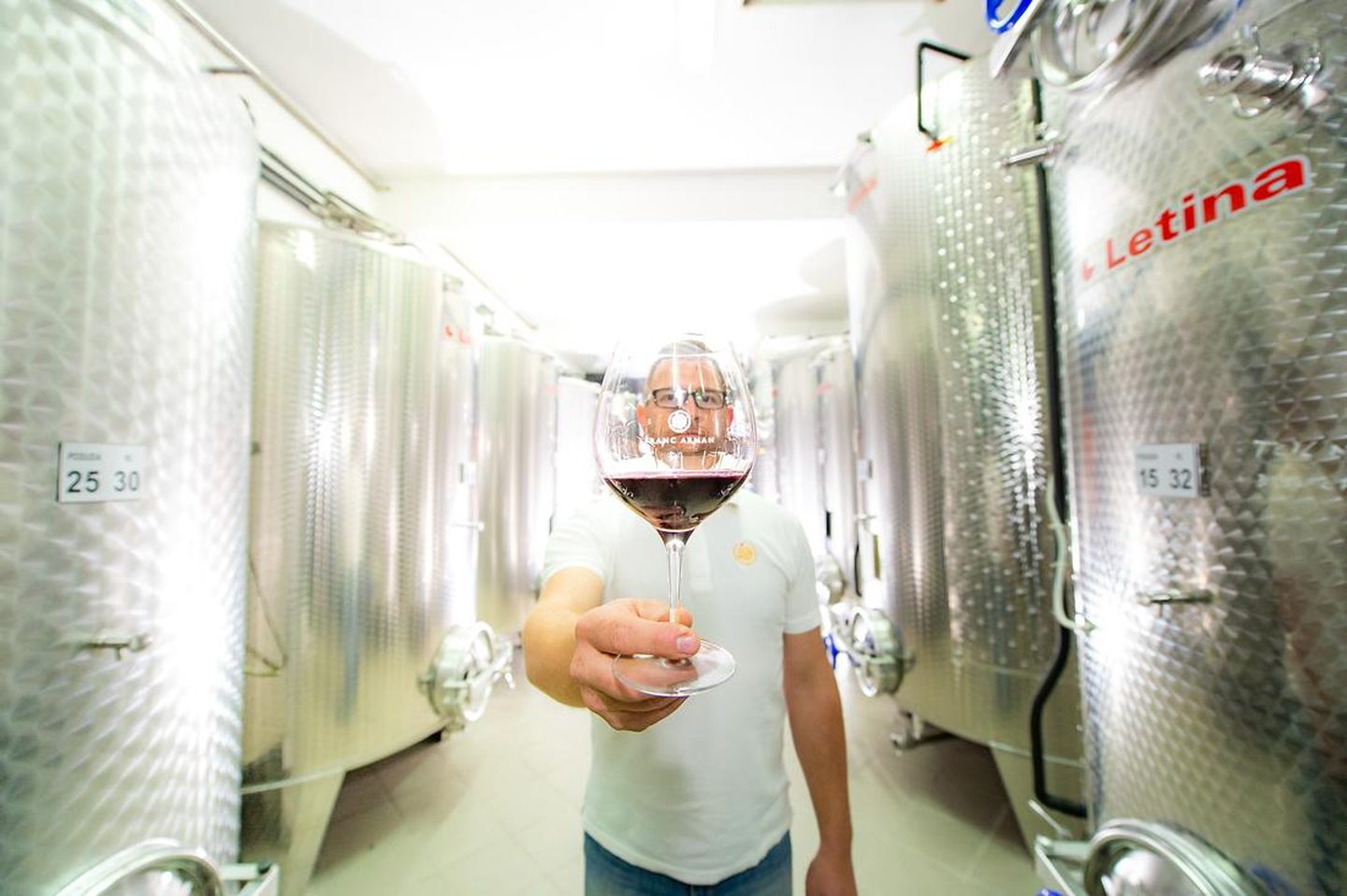 Winemaker between stainless steel tanks holding a glass of red wine with the Franc Arman logo toward the camera