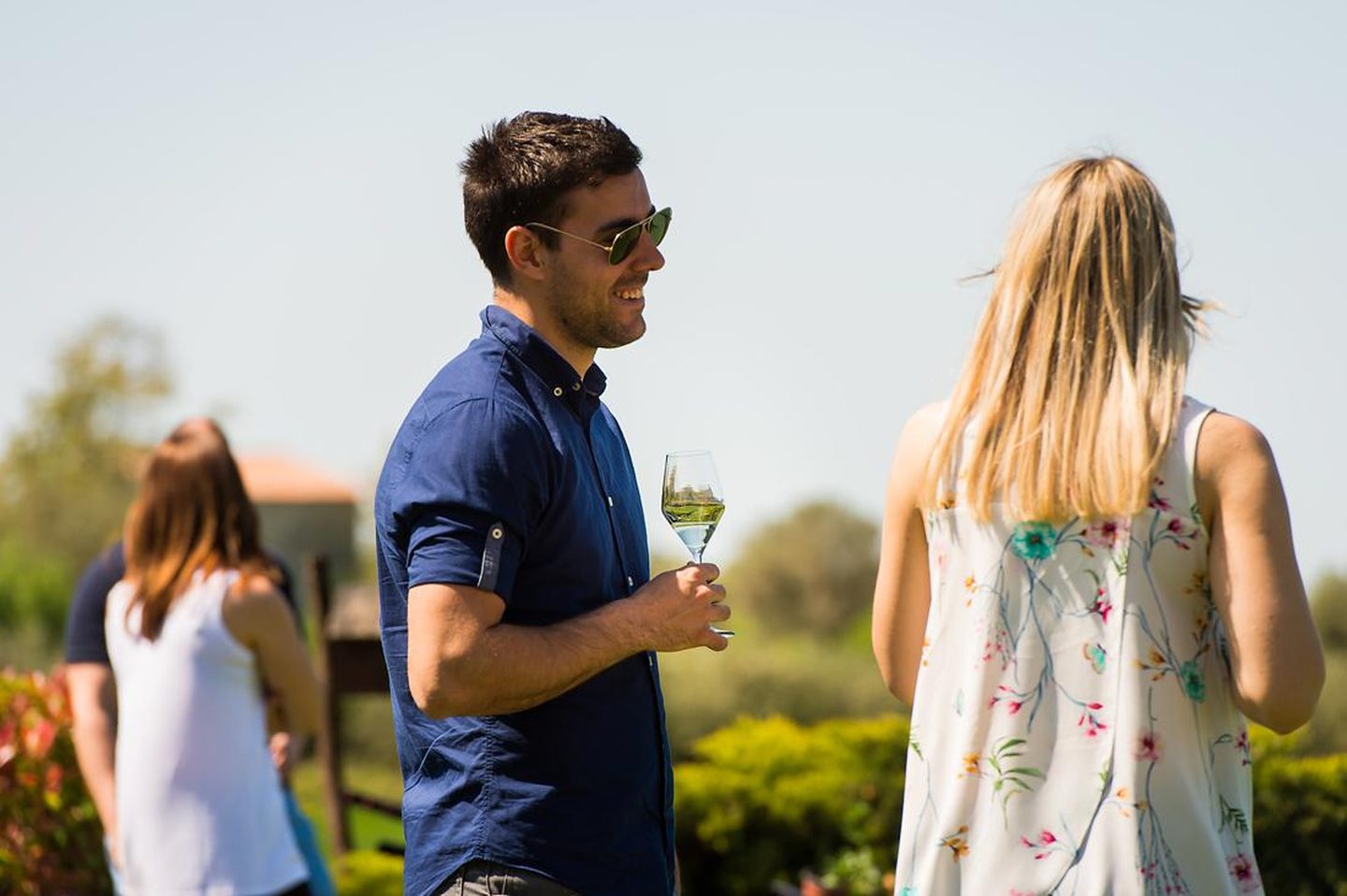 Guest in sunglasses holding a glass of white wine outdoors among other visitors on the estate lawn