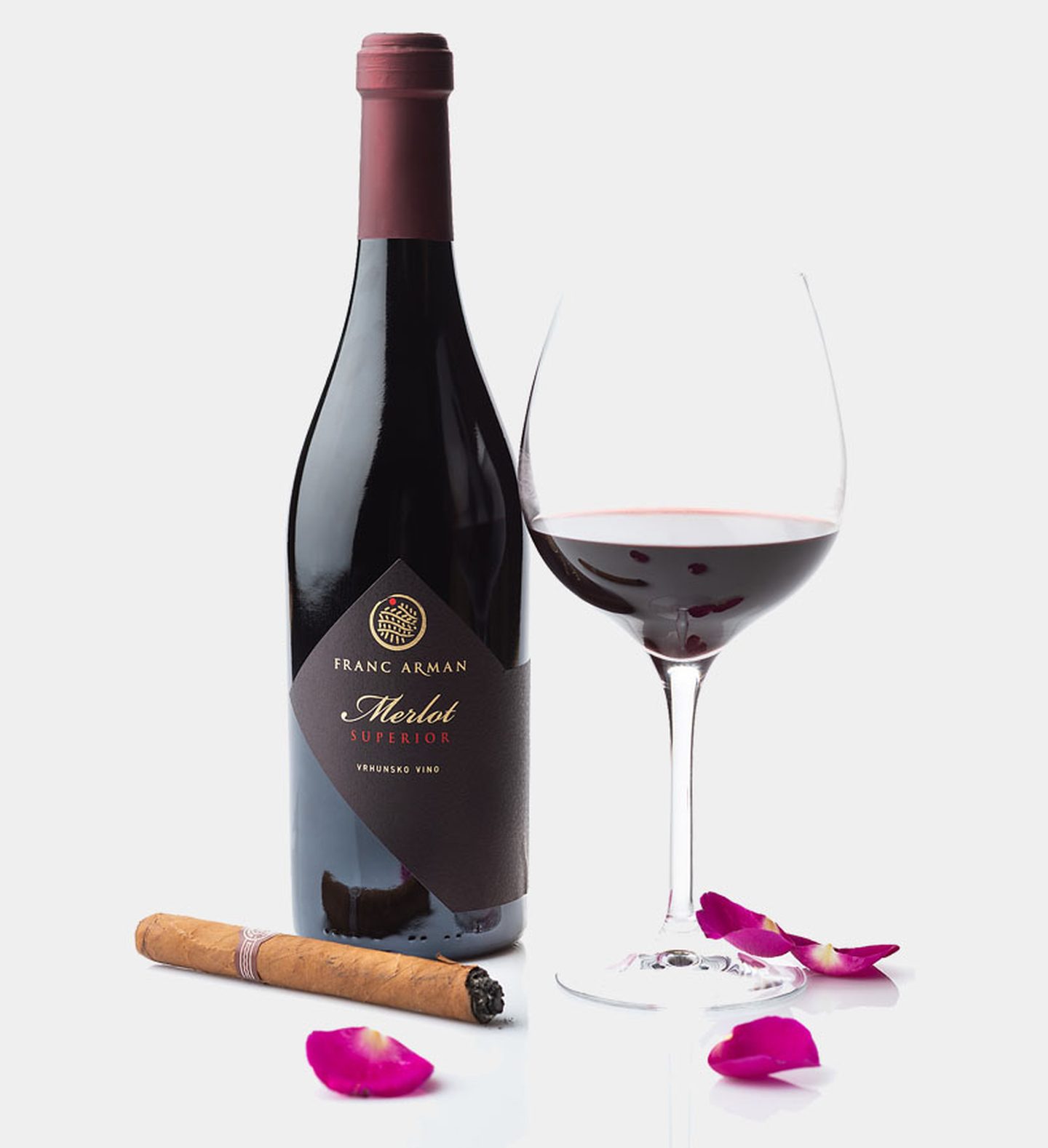 Bottle of Franc Arman Merlot Superior with kite label, a glass of red wine, a cigar and rose petals on white
